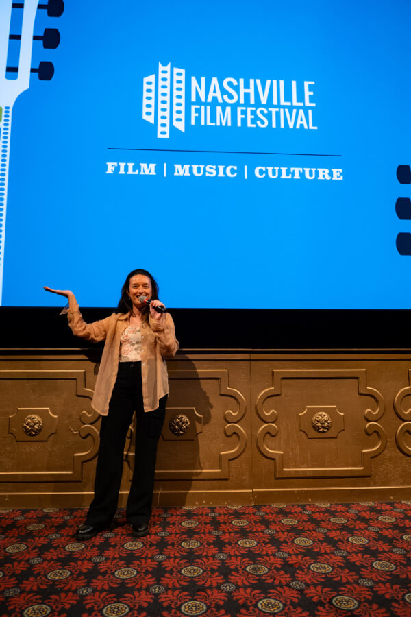 A person stands on stage holding a microphone, presenting in front of a large screen displaying "Nashville Film Festival" along with the words "Film | Music | Culture."