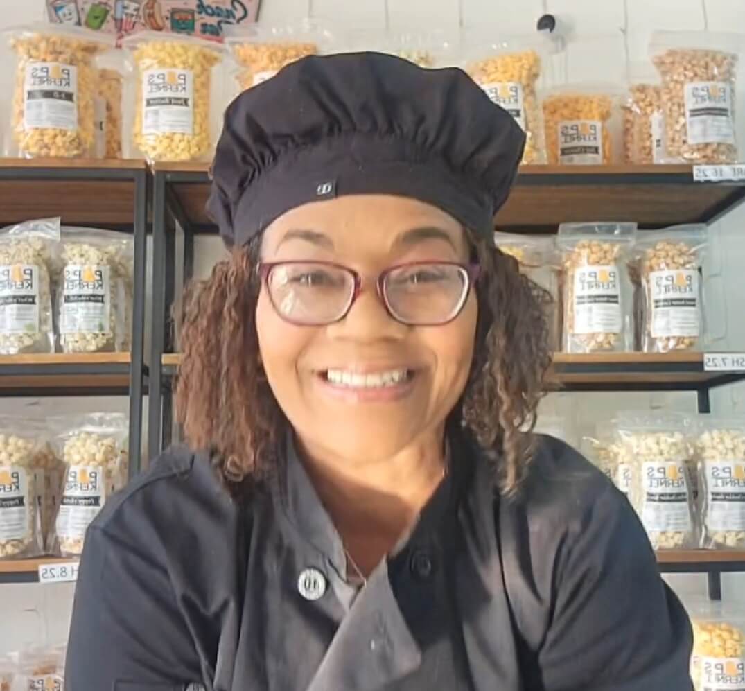 A person wearing glasses and a black chef's uniform is smiling, standing in front of shelves filled with bags of popcorn.
