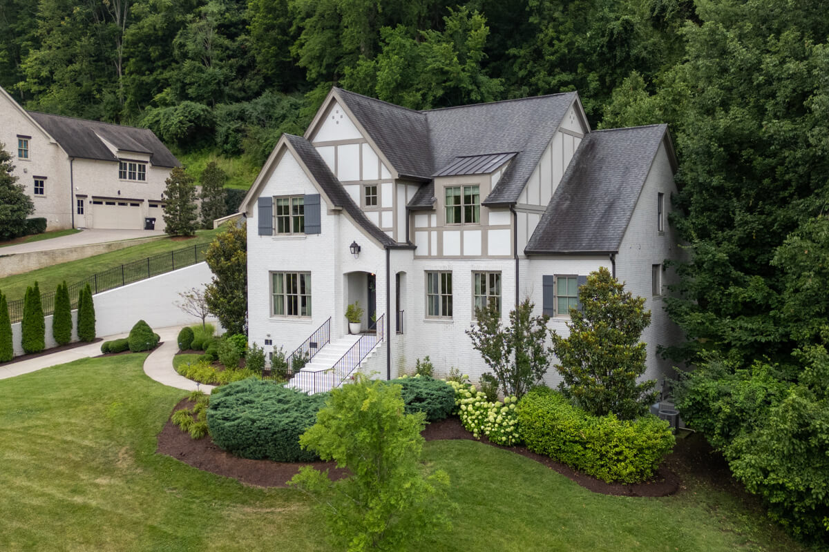 A two-story white Tudor-style house with dark roof accents, surrounded by landscaped greenery, sits adjacent to a driveway and lawn at 1049 Stockett Dr, backed by a dense forest in Franklin, TN.