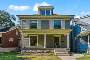 A two-story house on Bassett Avenue, with a front porch, gray siding, and a neatly manicured yard, sits elegantly under the clear blue sky in Louisville, KY.
