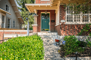 The front entrance of this red brick house at 2521 Ransdell Avenue in Louisville boasts a green door, surrounded by lush greenery and vibrant flowers. Steps lead to the door, and a security sign is subtly placed in the garden.