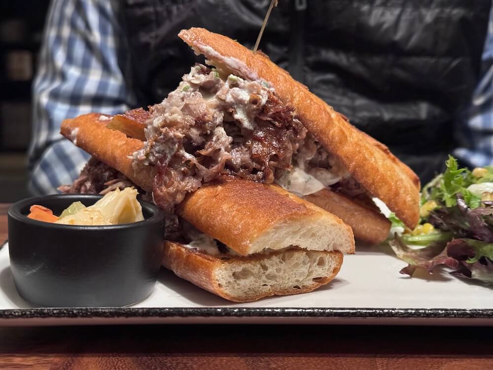 A hearty sandwich with shredded meat on toasted grain bread is served with a side of pickled vegetables and a delicious fresh salad.