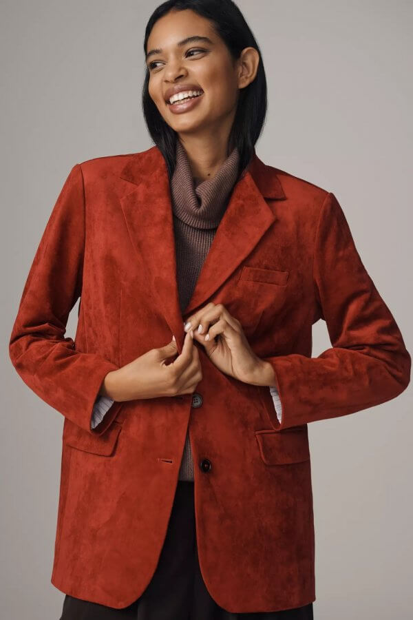 A person smiles while wearing a rust-colored blazer, the perfect piece of outerwear for fall fashion, over a brown turtleneck.