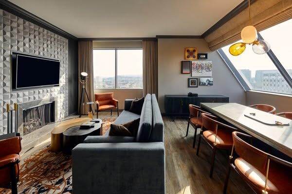Modern living room in Midtown with a gray sofa, textured walls, a flat-screen TV, and a cozy fireplace. The space features a dining table with vibrant orange chairs and large windows offering views reminiscent of Luxe Hotels in Nashville, complemented by geometric art on the walls.