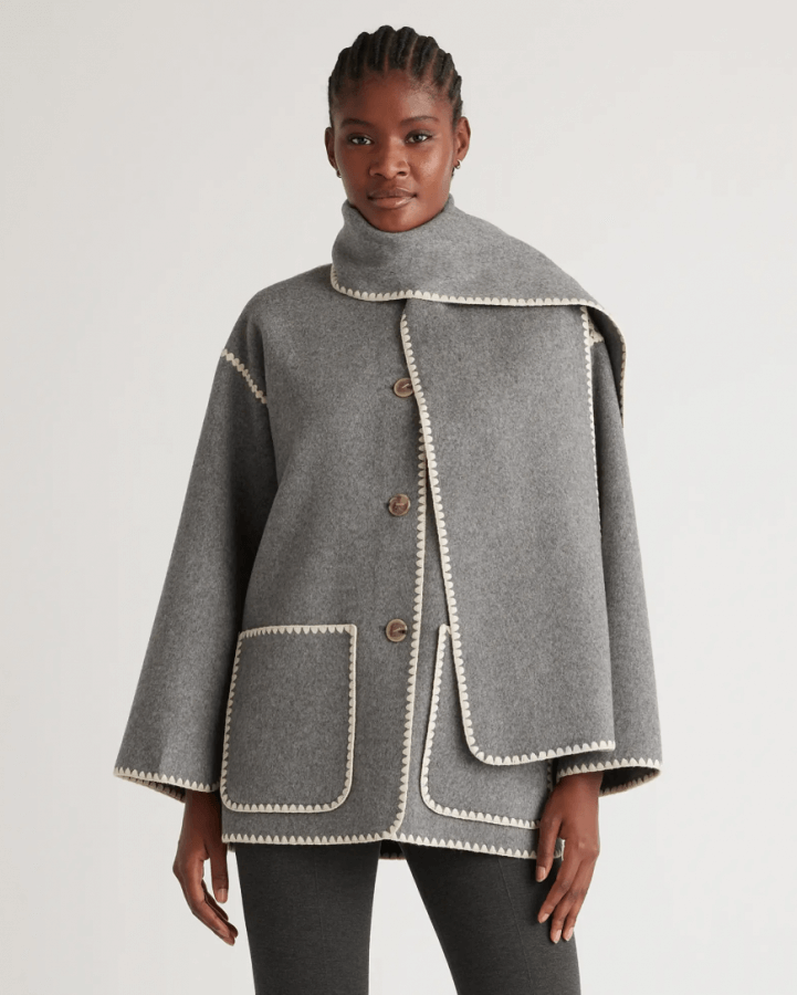 A person wearing a grey coat with white trim and buttons stands against a plain background, showcasing one of this season's stylish fall jackets.