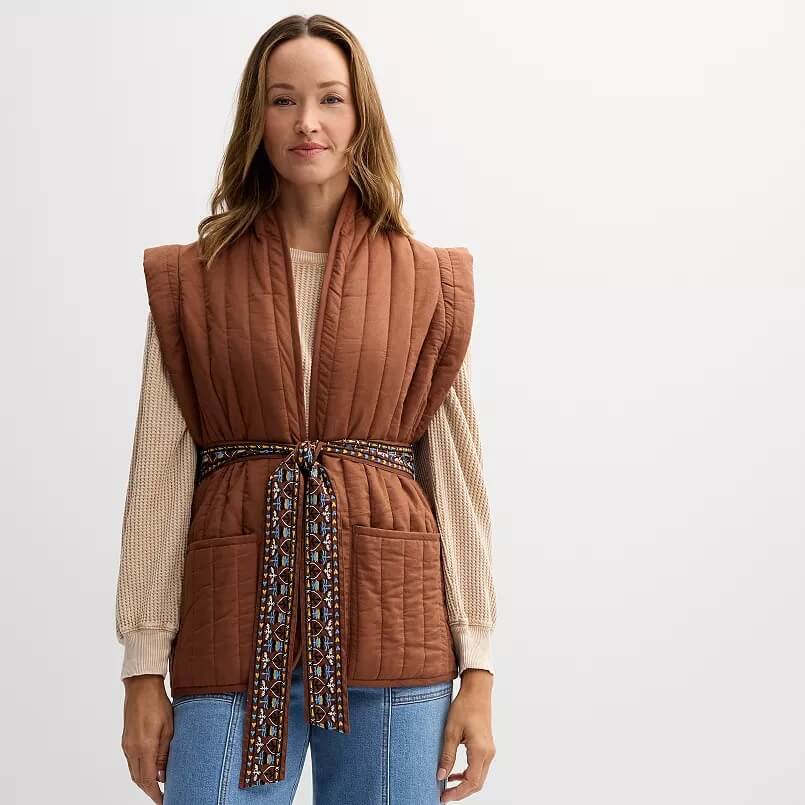 A person stands against a plain background, showcasing a brown quilted vest reminiscent of trendy fall jackets, paired with a patterned belt over a beige sweater and blue jeans.