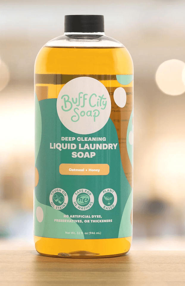 A bottle of Buff City Soap Deep Cleaning Liquid Laundry Soap with oatmeal and honey. The label also states it's tough on stains, safe for HE washers, and plant-based with no artificial additives.