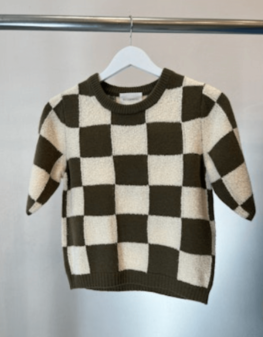A short-sleeved, checkered sweater in black and white hangs on a white hanger against a plain background, perfect for crisp October days.