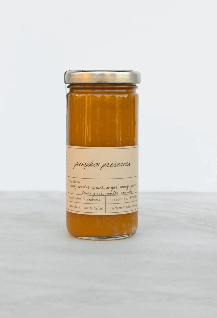 A glass jar with a metal lid containing pumpkin preserves. The label lists ingredients and notes that it is handmade in a small batch.