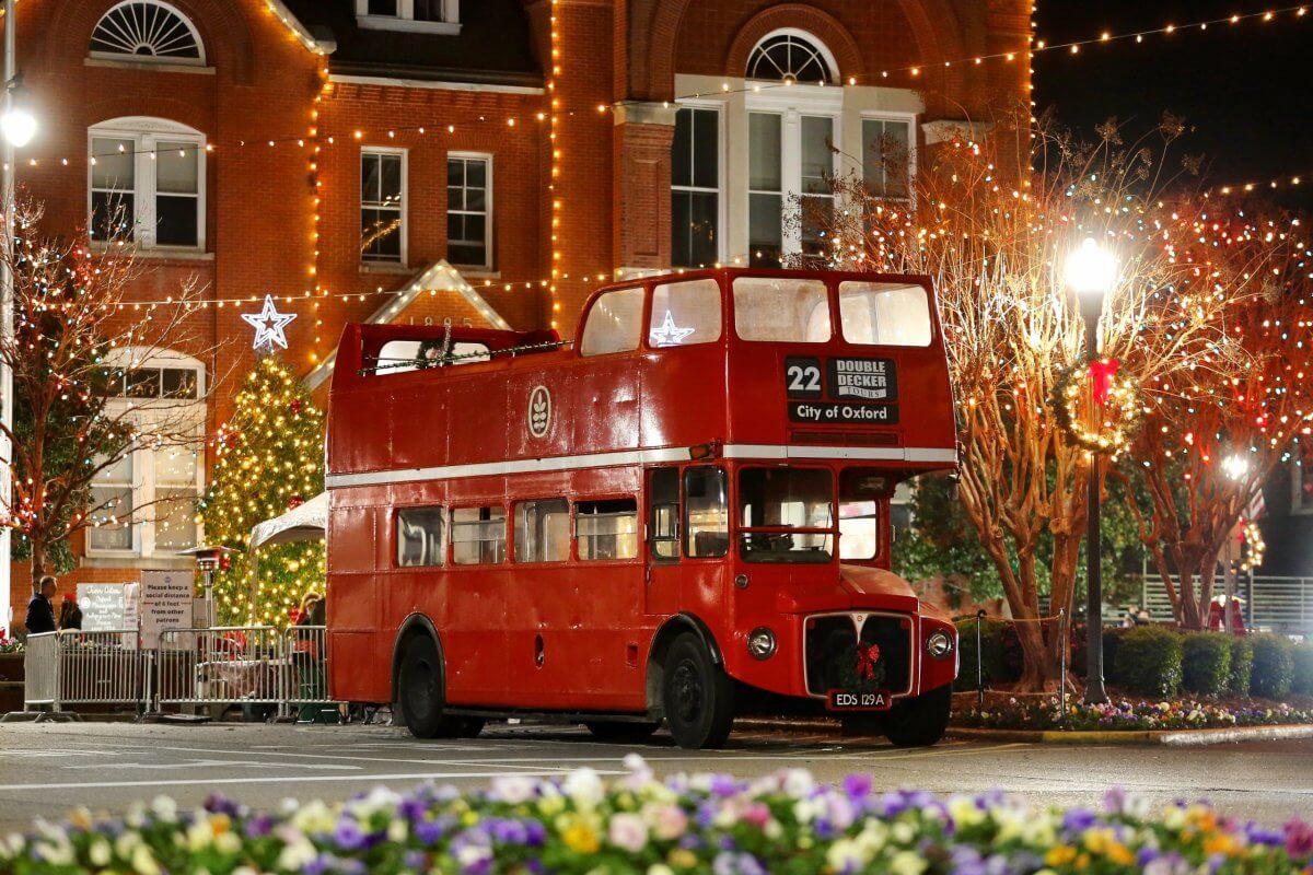 A classic red double-decker bus, adorned with festive lights, is parked on a street. In the background, a brick building and Christmas tree evoke the charm of holidays in one of Mississippi's best towns.