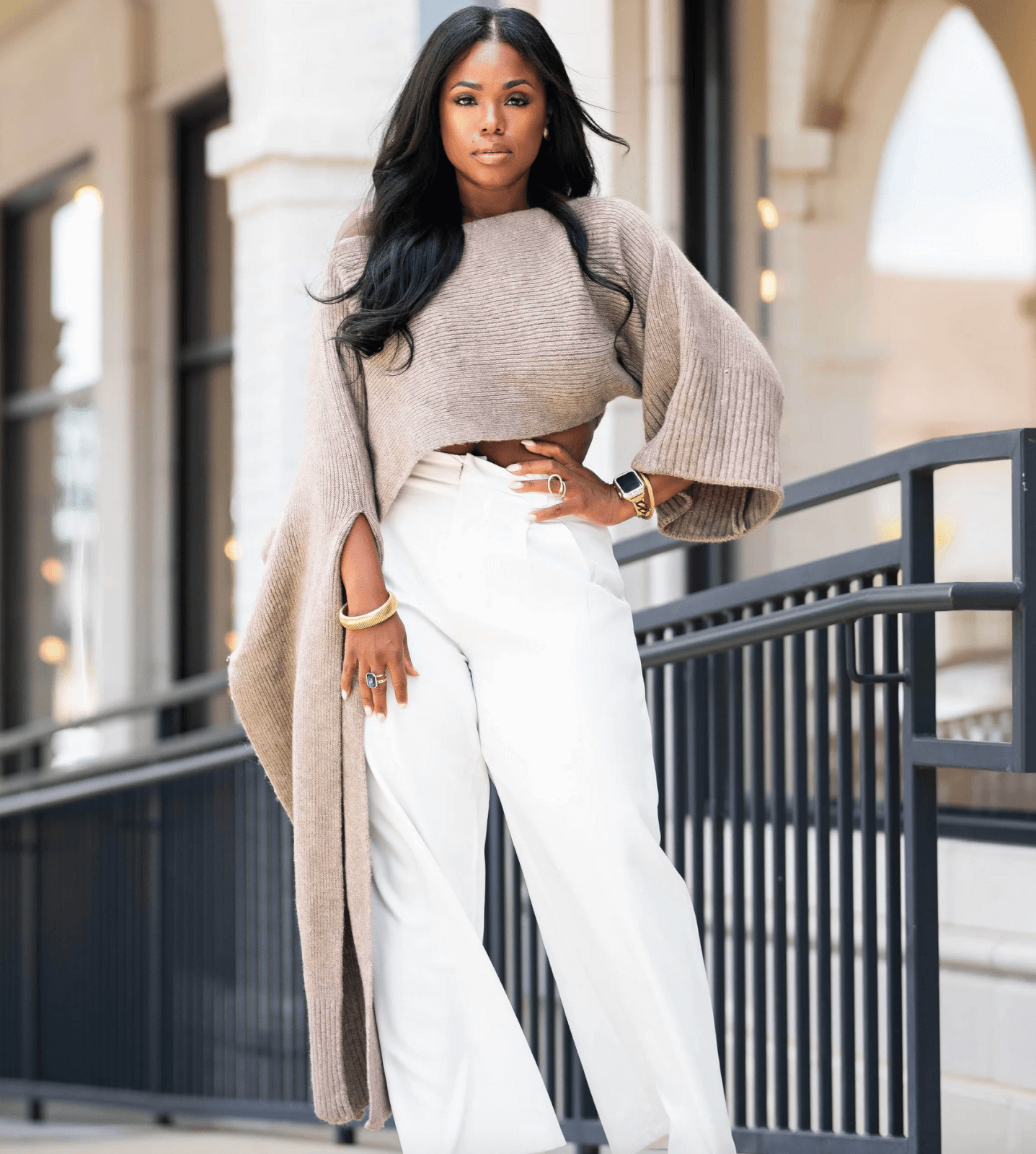 Woman in a taupe sweater and white pants leans on a black railing, with beige building in the background.