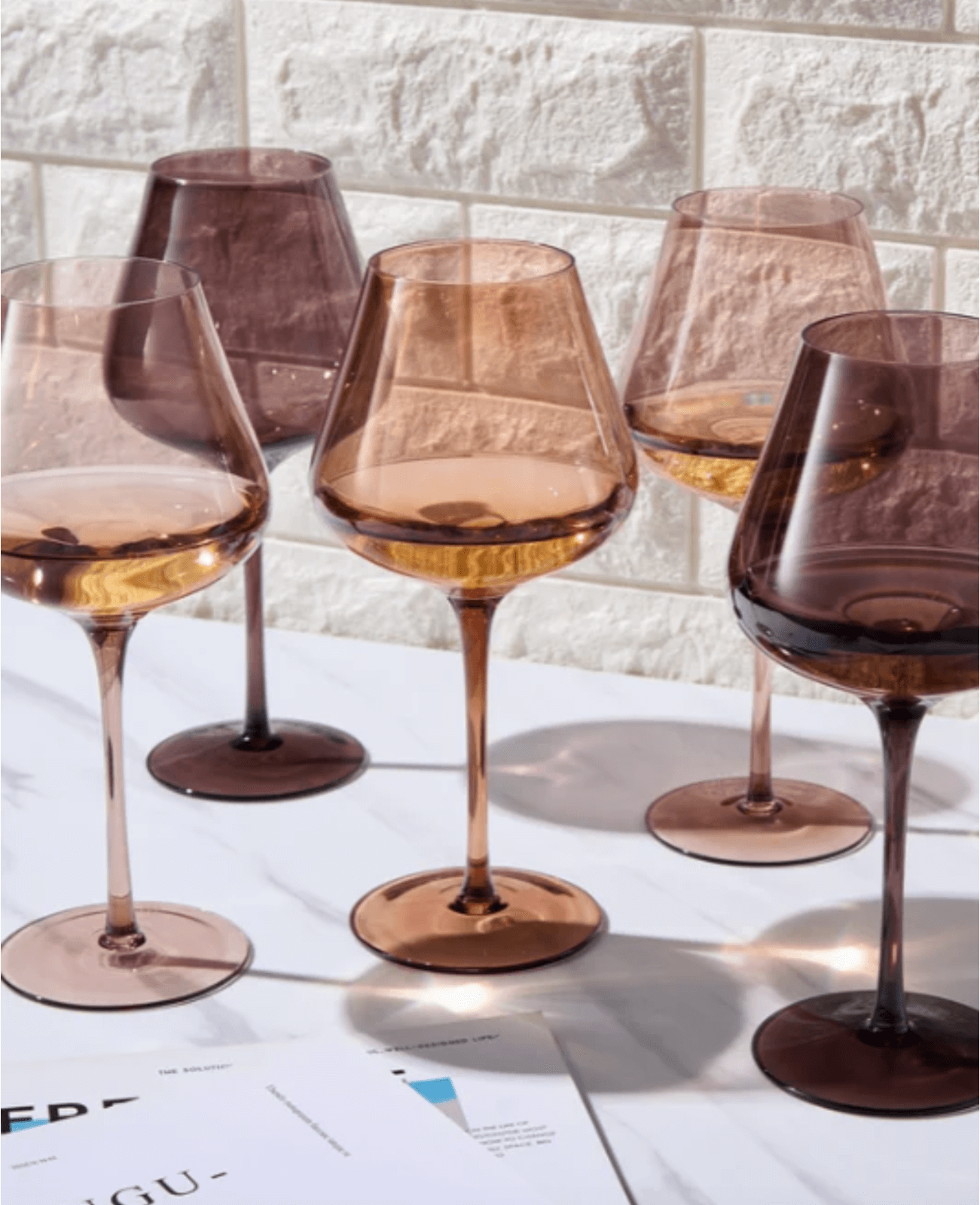 Five tinted wine glasses with thin stems sit on a marble surface, casting shadows. A stack of papers is partially visible in the foreground.