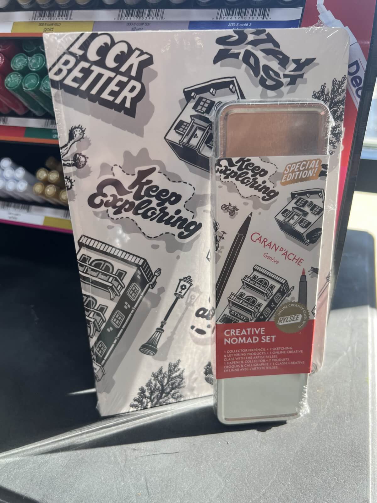 A special edition Caran d'Ache Creative Nomad Set is displayed on a store shelf next to a notepad with "Keep Exploring" printed on the cover.
