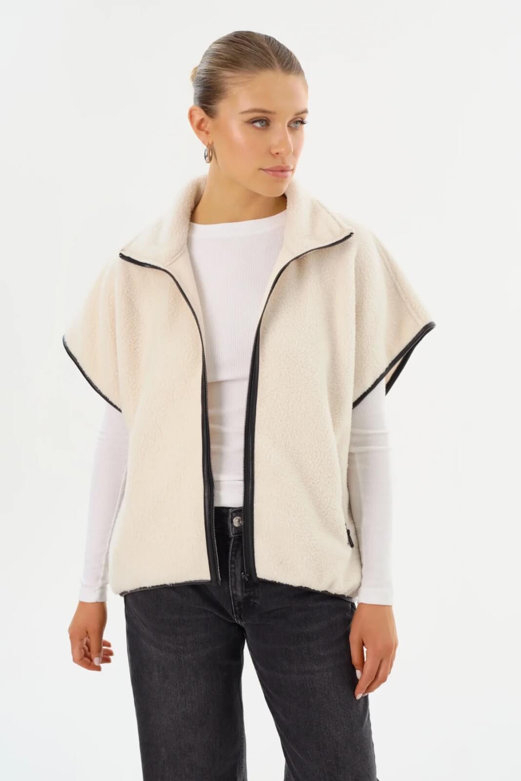 Against a plain backdrop, a woman wears a white zip-up vest over a long-sleeve shirt and black jeans.
