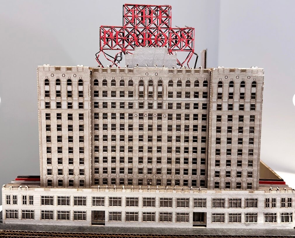 A scale model of a multi-story building features a detailed facade adorned with a striking red lattice structure on the roof.