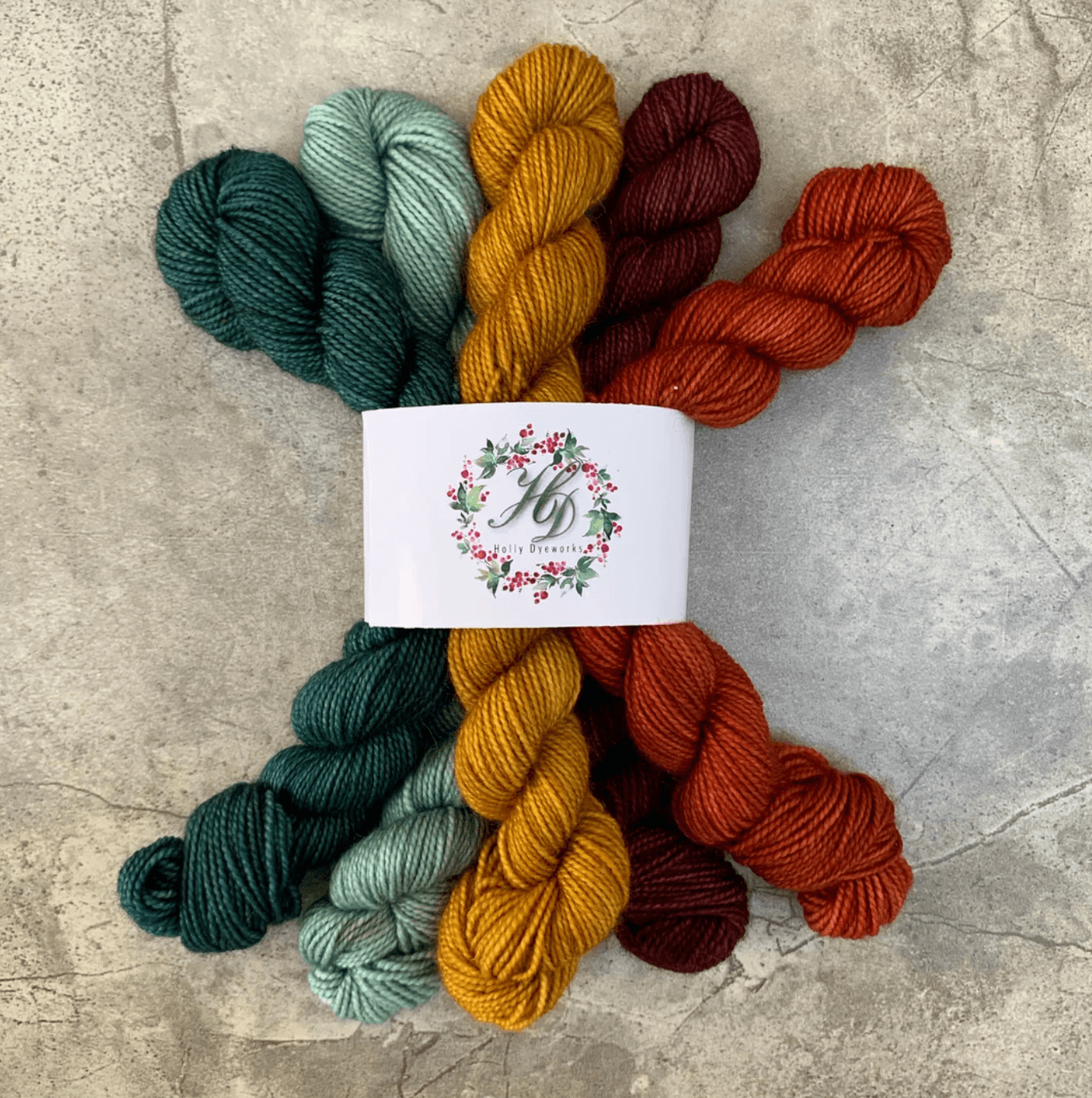 Five skeins of yarn in green, teal, yellow, orange, and red, arranged in a fan shape with a floral logo label in the center, on a textured gray surface.