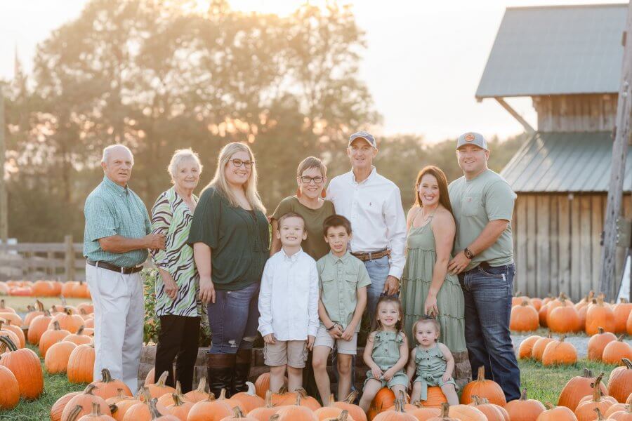 A group of nine people, including adults and children, stands in a pumpkin patch with a barn in the background. The scene, set during daylight in one of Nashville's charming pumpkin patches, captures the essence of fall October 2024.