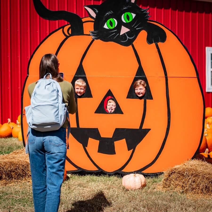 A lively scene captures children peeking through a large pumpkin cutout, adorned with a curious black cat, set against a vibrant red wall. Surrounded by hay bales and pumpkins, this enchanting moment echoes the charm of Nashville's pumpkin patches in the fall.