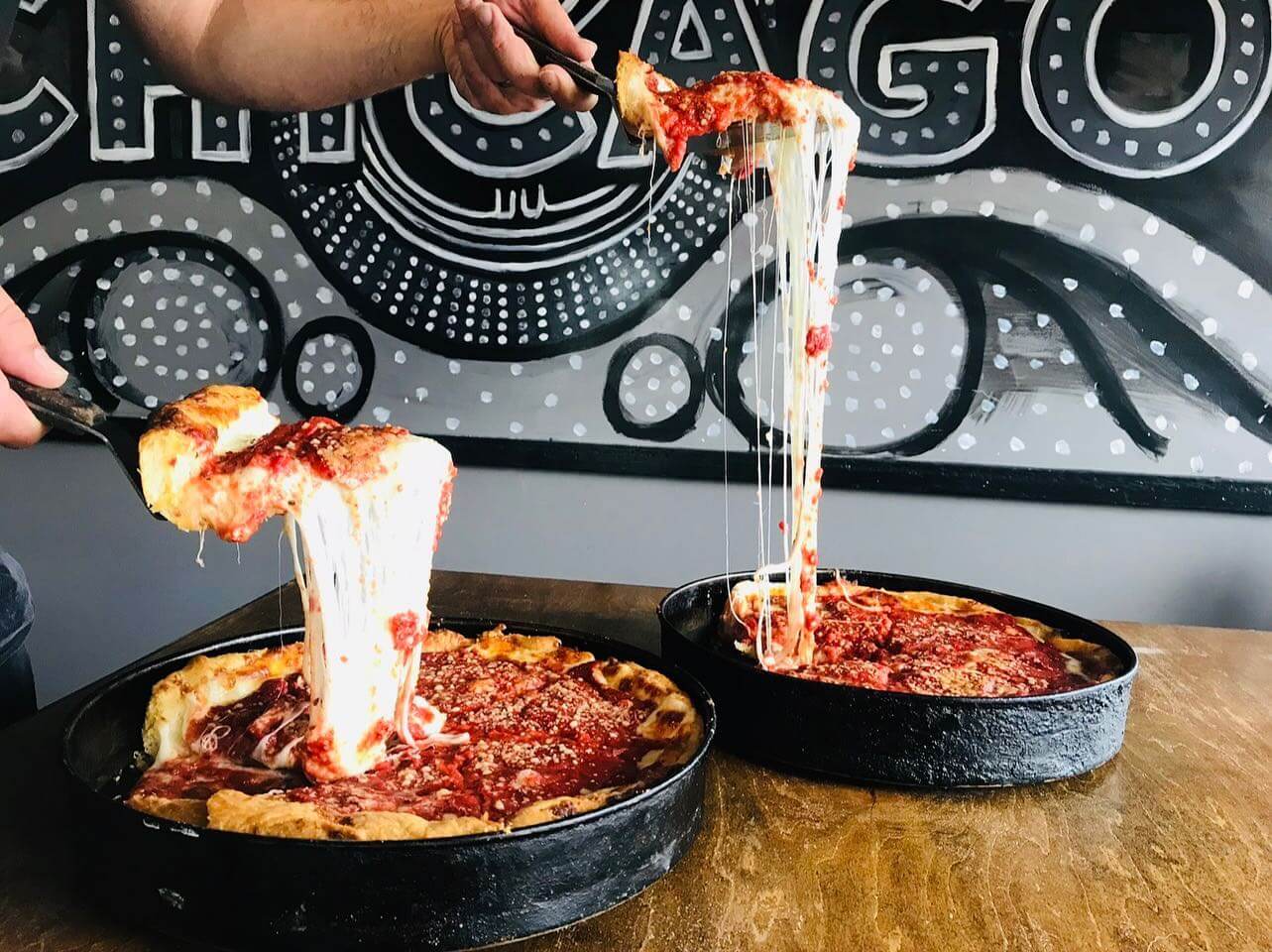 Two Chicago-style deep-dish pizzas with melted cheese being pulled from slices over a wooden table, a decorative "Chicago" sign in the background, perfect for an NFL watch party.