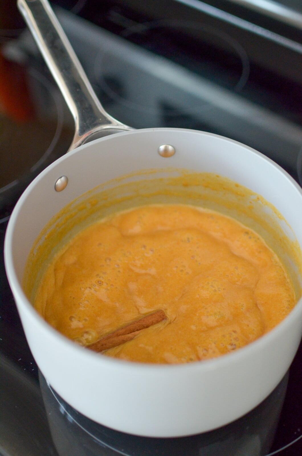 A white saucepan on the stove holds a bubbling orange mixture with a cinnamon stick, perfect for crafting homemade pumpkin syrup.