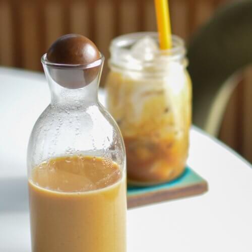 A glass bottle with a round stopper contains a brown liquid, sitting on a table. In the background, a mason jar with a yellow straw holds a creamy iced drink.