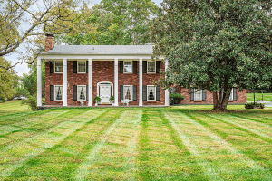 A red-brick, two-story house with white columns and black shutters sits gracefully at 6104 Innes Trace Road, Louisville KY, behind a neatly mowed lawn, surrounded by trees.
