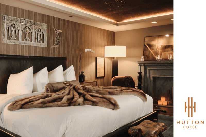 Experience luxury at its finest in this Nashville hotel room featuring a large bed draped with a fur blanket, exquisite wall art, and a cozy wooden accent wall. Unwind by the warm fireplace amidst ambient lighting, all with the elegant Hutton Hotel logo proudly displayed.