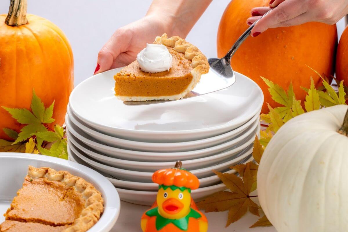A hand serves a slice of pumpkin pie with whipped cream onto a stack of plates, surrounded by pumpkins, autumn leaves, and a toy duck—perfect inspiration when searching for Memphis restaurants open on Thanksgiving.