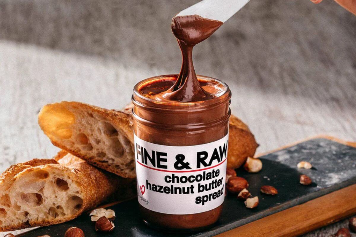 A jar of Fine & Raw chocolate hazelnut butter spread with a knife lifting some spread, sliced bread, and hazelnuts on a wooden board—an indulgent treat perfect for the Memphis Gift Guide.