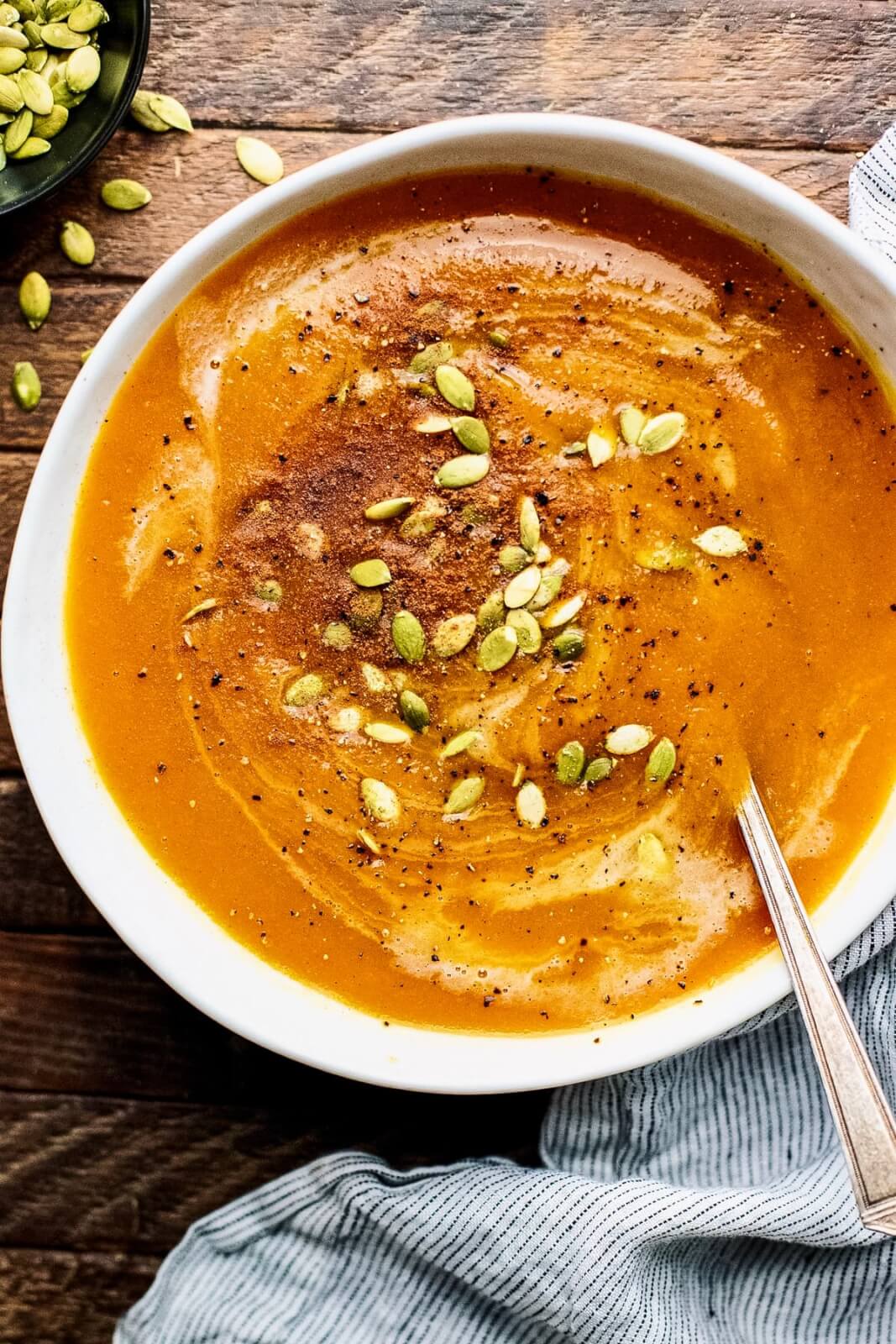 A bowl of creamy pumpkin soup topped with pumpkin seeds and black pepper, perfect for your Nashville Thanksgiving takeout feast, with a spoon on the side.
