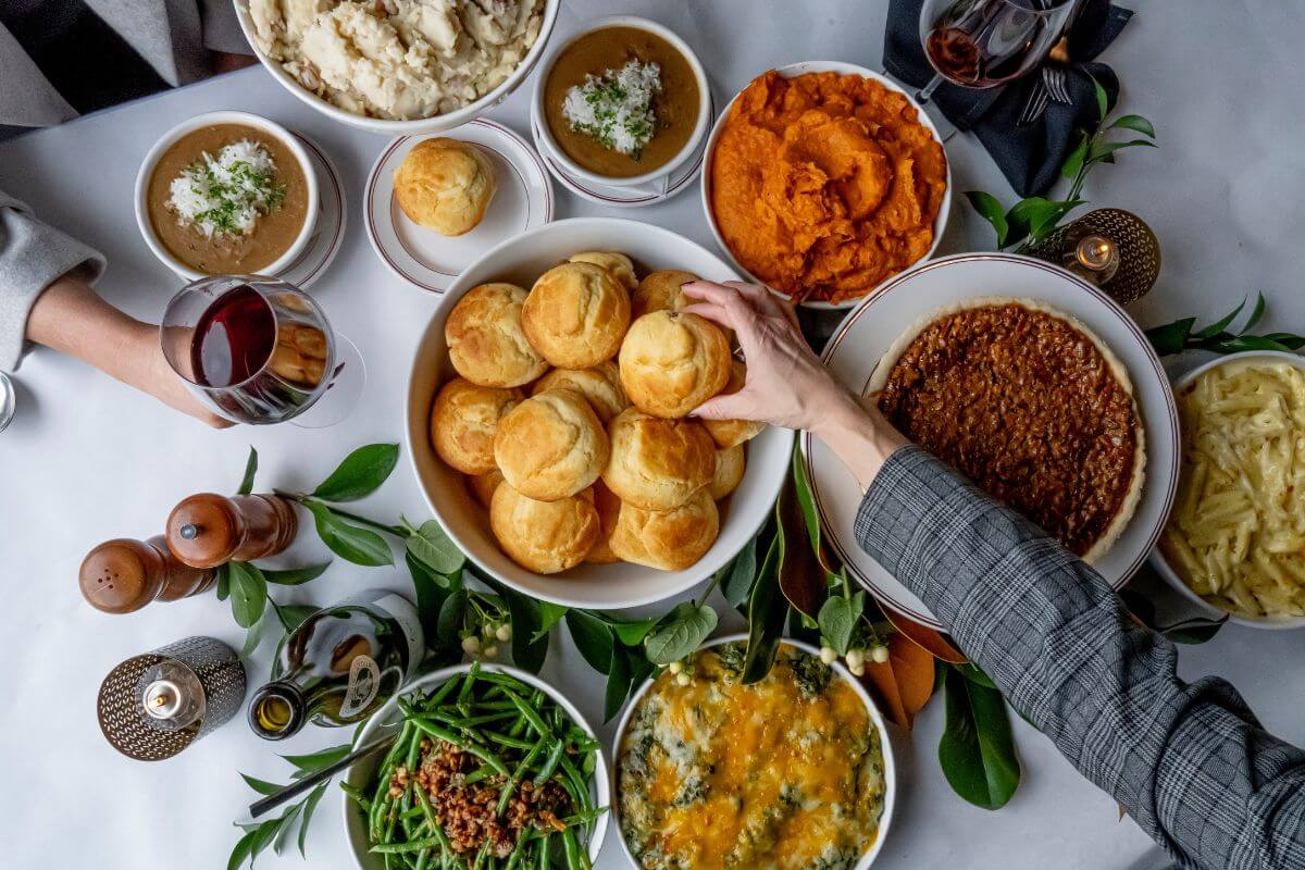 A table set for a Nashville Thanksgiving takeout feast, with dishes like biscuits, mashed potatoes, sweet potatoes, green beans, and casserole, as two people reach for food and glasses of red wine.