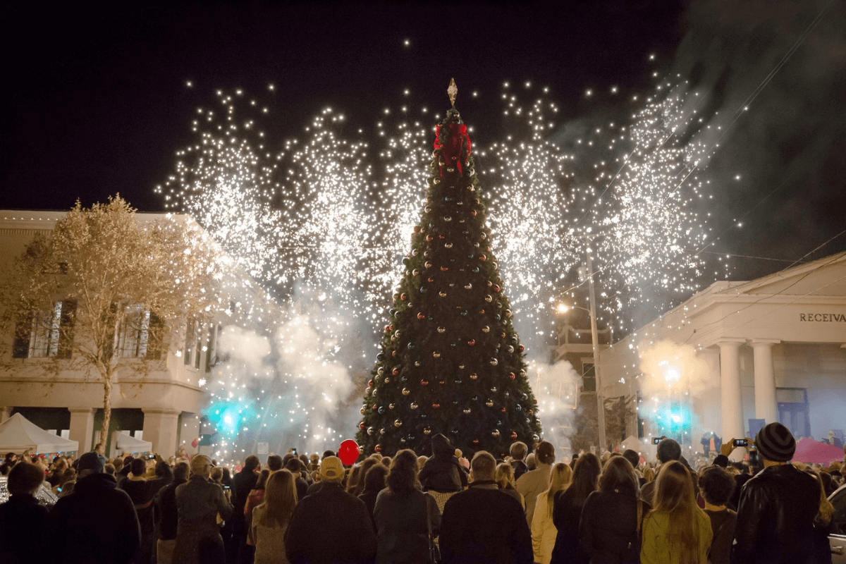 A large crowd gathers at night to watch fireworks erupting behind a decorated Christmas tree in a city square, celebrating one of the southern events that light up December.