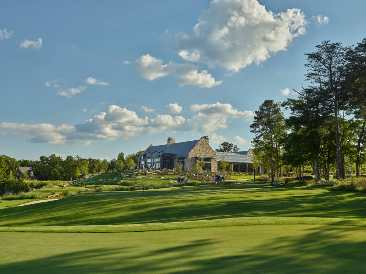 A large stone clubhouse sits beside a well-maintained golf course in one of the area's premier second home communities, under a partly cloudy sky and surrounded by lush trees and greenery.