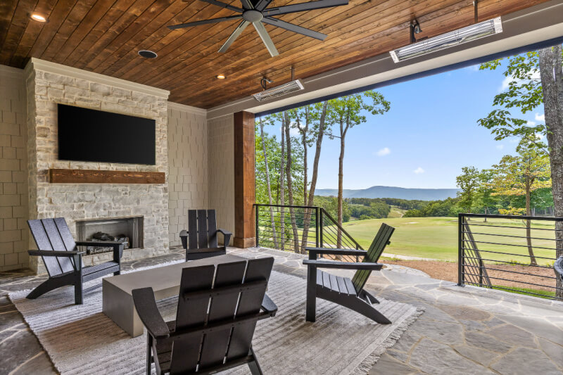 Covered patio with stone fireplace, wall-mounted TV, four black chairs, and a modern table—perfect for relaxing in second home communities while enjoying stunning views of trees and mountains in the background.