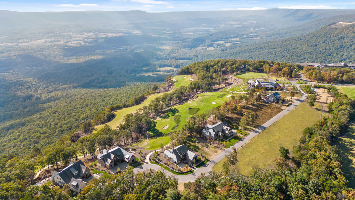 Aerial view of a golf course surrounded by forested hills, second home communities, residential houses, and club buildings under a partly cloudy sky.