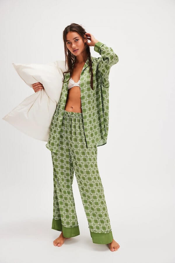 A person in festive green checkered pajamas holds a white pillow, standing barefoot in a room with a plain background.