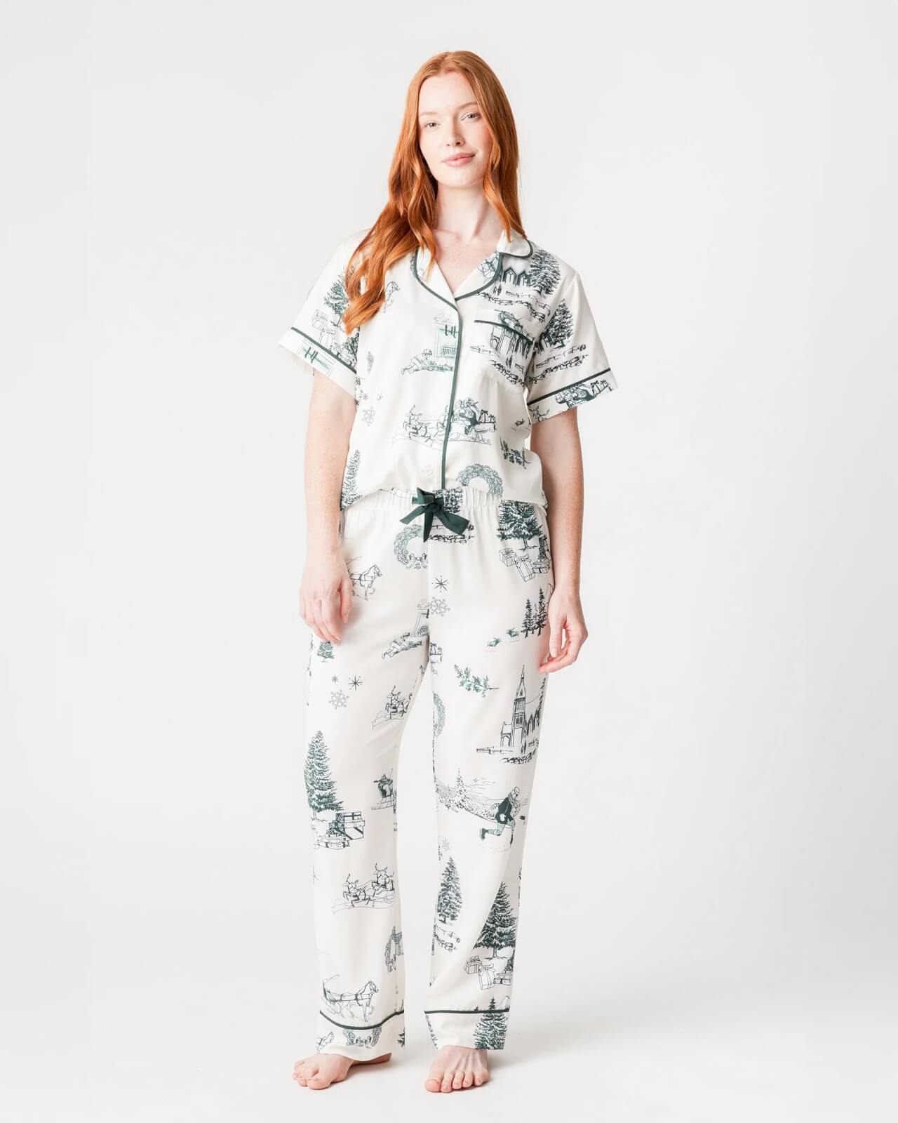 A person in holiday pajamas adorned with black illustrations stands barefoot against a plain background, ready to embrace the festive spirit.