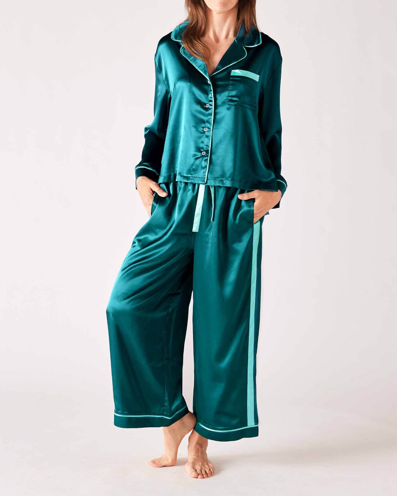 A person stands barefoot on a plain background, wearing teal satin pajamas with white trim.