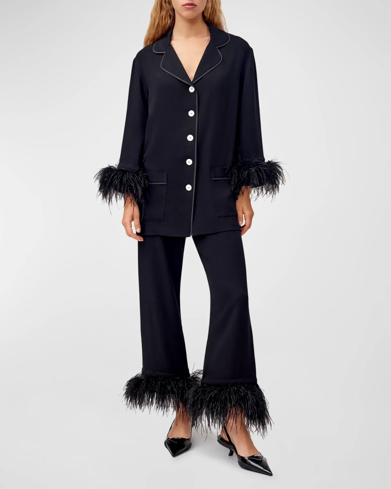 A person stands against a plain background, exuding holiday style in a black pajama set adorned with feather trim on the sleeves and pant legs.