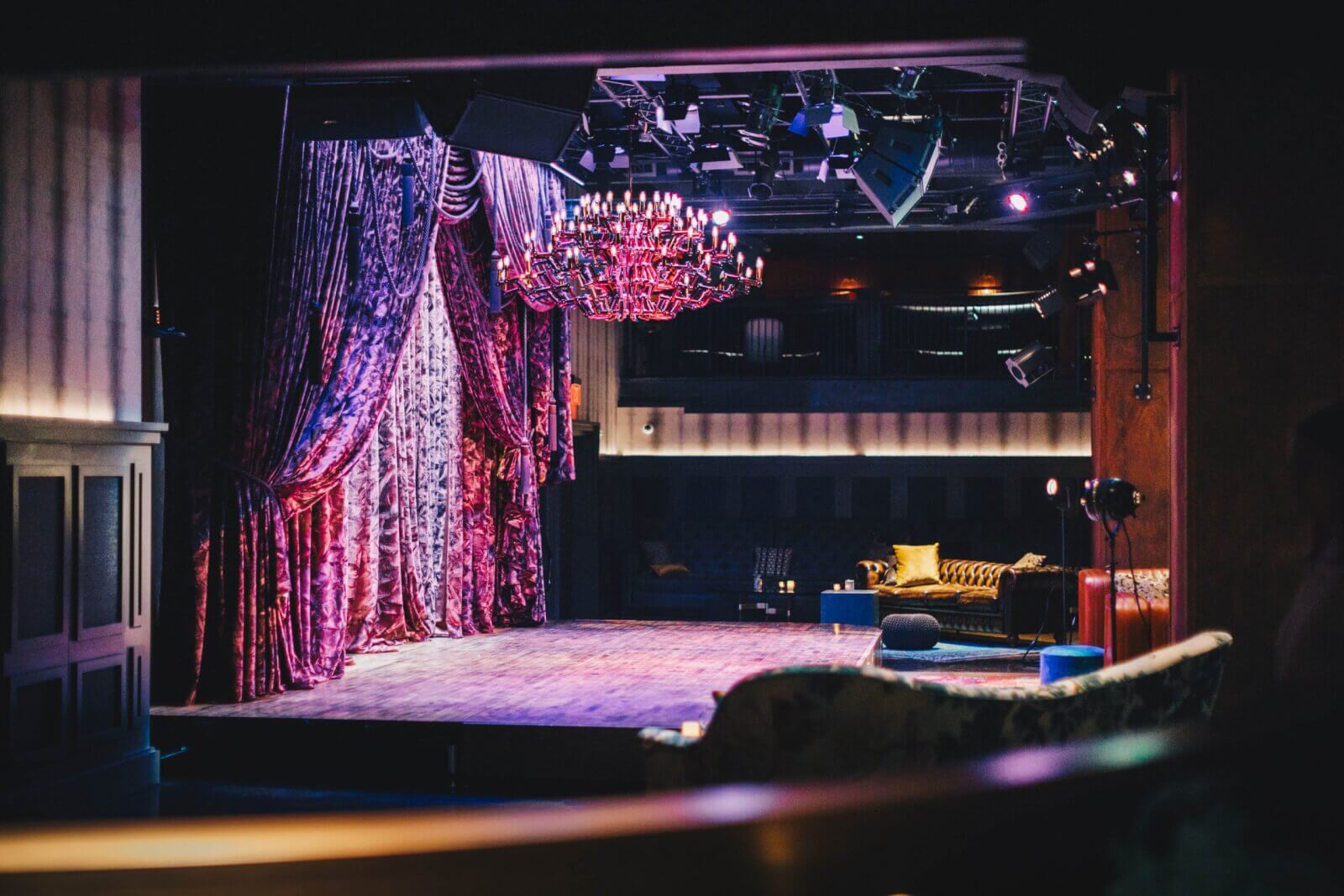 The empty stage of the Hutton Hotel, draped in velvet curtains and dimly lit by a grand chandelier, sets an intimate scene. In the background, cozy seating with plush sofas and cushions invites guests to relax in style.