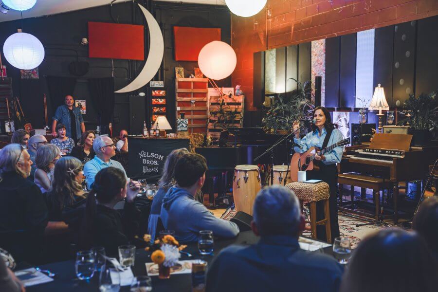 Meet Nashville's Studio Mama, Rebecca Wood - 3 Lori McKenna performs with a guitar in a cozy, dimly lit venue. The audience sits at tables, captivated by her performance. Lights and a crescent moon decoration set a warm atmosphere.