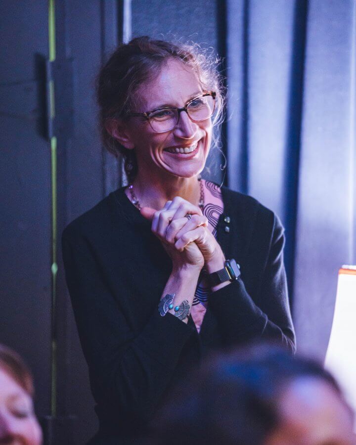 Meet Nashville's Studio Mama, Rebecca Wood - 5 Rebecca Wood smiles warmly with glasses. Her hands are clasped gracefully against a dimly lit background, capturing both charm and mystery in the moment.