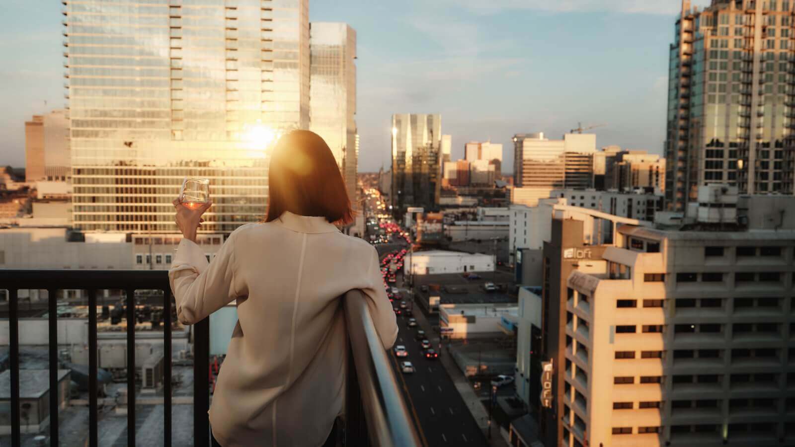 A person leans on a balcony railing at the Hutton Hotel, holding a drink and gazing at the cityscape during sunset.
