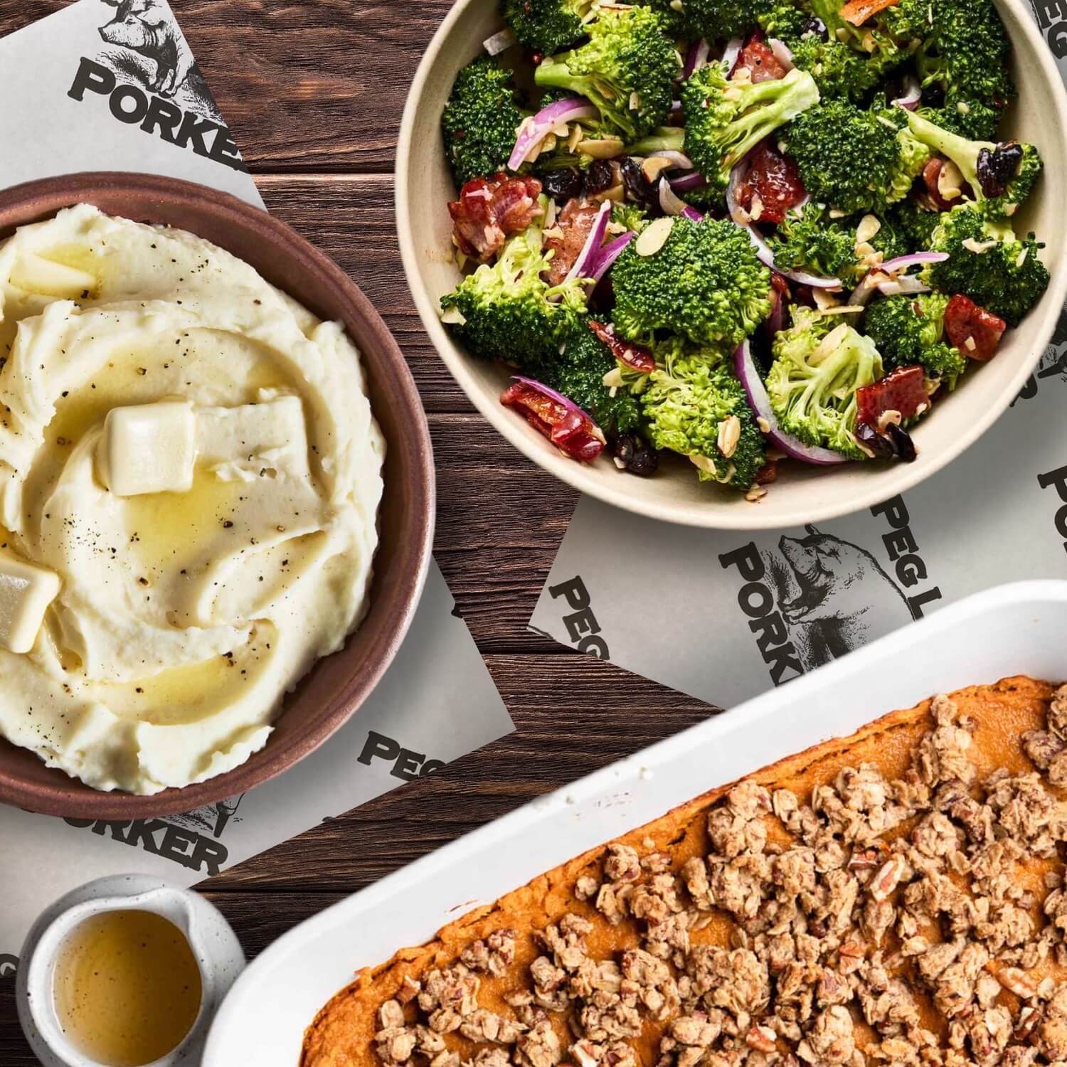 Bowls of mashed potatoes and broccoli salad alongside a glass dish of crumble topping and a small cup of sauce, set on a wooden table—perfect for your Nashville Thanksgiving takeout spread.