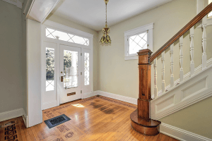 The entrance hall at 1316 Everett Avenue in Louisville boasts a wooden staircase, decorative moldings, and a brass chandelier. A white front door with glass panels leads outside, while the light wood floor showcases intricate patterned inlays.
