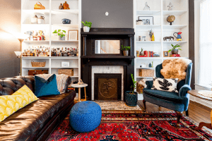 Nestled in a home on Highland Avenue in Louisville, KY, this cozy living room features a leather sofa, blue armchair, and ornate fireplace. Shelves brim with bottles and books, while a red patterned rug unifies the charming space.