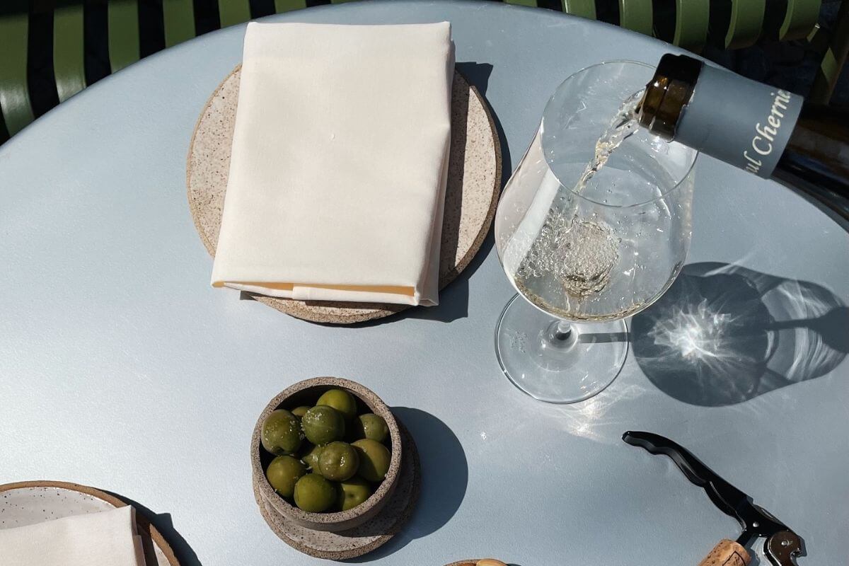 A bottle pours white wine into a glass on a pale blue table next to a bowl of green olives, a folded napkin, and a wine opener—perfect for enjoying Birmingham gift experiences.