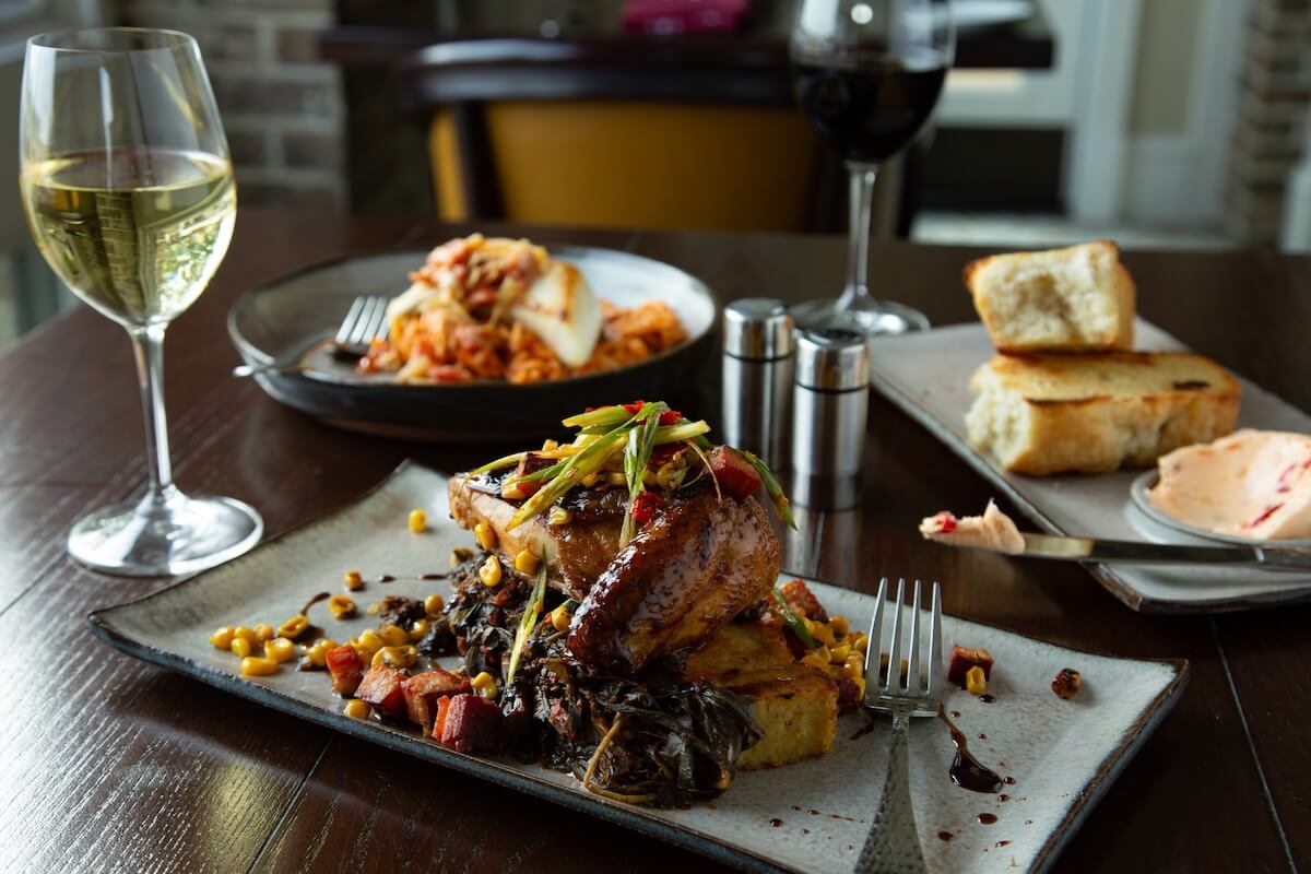 A plated gourmet meal with roasted meat, vegetables, and sauce, accompanied by a glass of white wine, bread, butter, and a pasta dish on a wooden table—perfect inspiration for Birmingham restaurants open on Christmas.