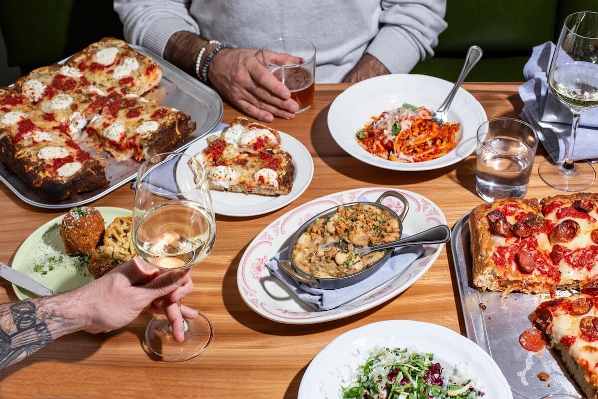 A wooden table set with Detroit-style pizza, pasta, garlic bread, salad, and wine welcomes guests at one of the Birmingham restaurants open on New Year's Day, with two people sitting and their hands visible.