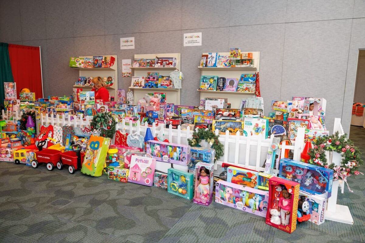 A display of assorted children's toys, arranged on shelves and behind a white picket fence at an indoor event, reminiscent of Birmingham toy drives.